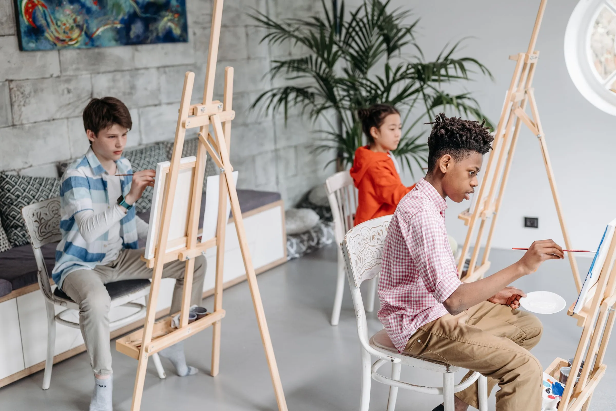 Three teenagers painting at wooden easels in a bright white studio, each turned to their own canvas