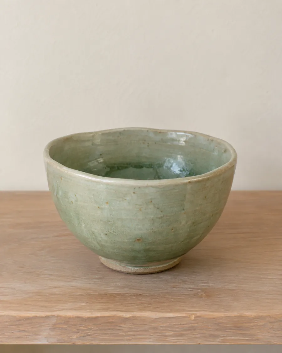 A wheel-thrown stoneware bowl with a pale green celadon glaze and a slightly wavy rim, on a wooden shelf