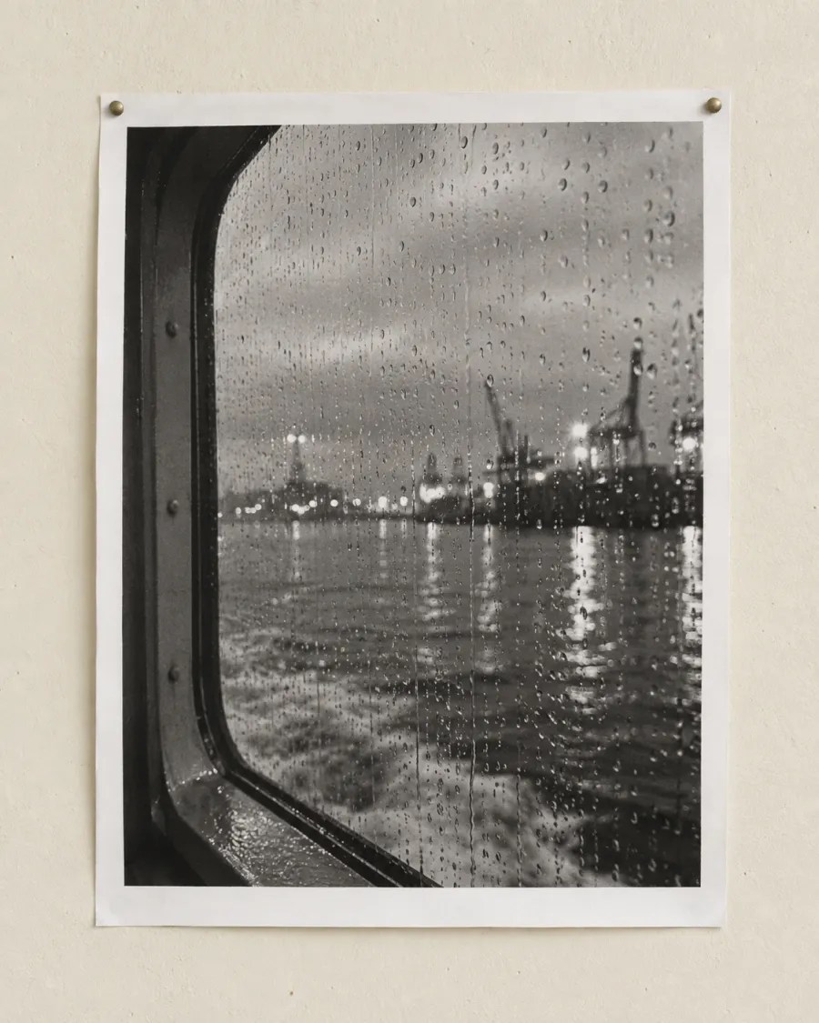 A black-and-white photograph of rain on a ferry window, harbour cranes and lights blurred behind the glass