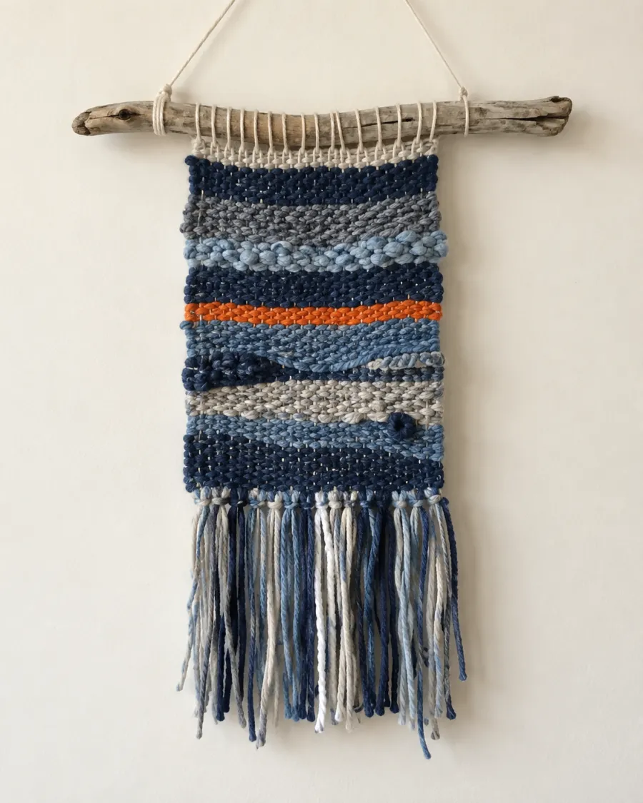 A small woven wall hanging in blues and greys with one orange stripe, hung from a piece of driftwood
